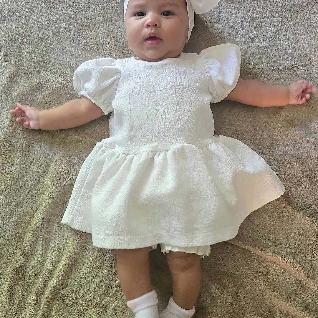 Christin joined the competition — help win amazing prizes! arms_outstretched, baby, blanket, bow, candid, child, curious_expression, cute, floor, headband, indoor, infant, lying_down, person, portrait, red_toy, socks, soft_texture, toy, white_dress