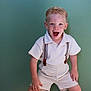 Giovanni participe au concours pour gagner de l'argent avec cette photo : child, boy, blond_hair, blue_eyes, suspenders, shorts, shirt, smiling, happy, portrait, indoor, floor, wooden_floor, green_wall, playful, cute, toddler, person, standing, sock