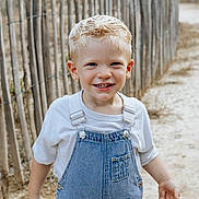 Giovanni participe au concours pour gagner de l'argent avec cette photo : child, boy, blonde_hair, blue_eyes, denim_overalls, white_tshirt, smiling, outdoor, fence, sand, path, portrait, happy, casual_clothing, daylight, nature, person, cute, young_child, summer