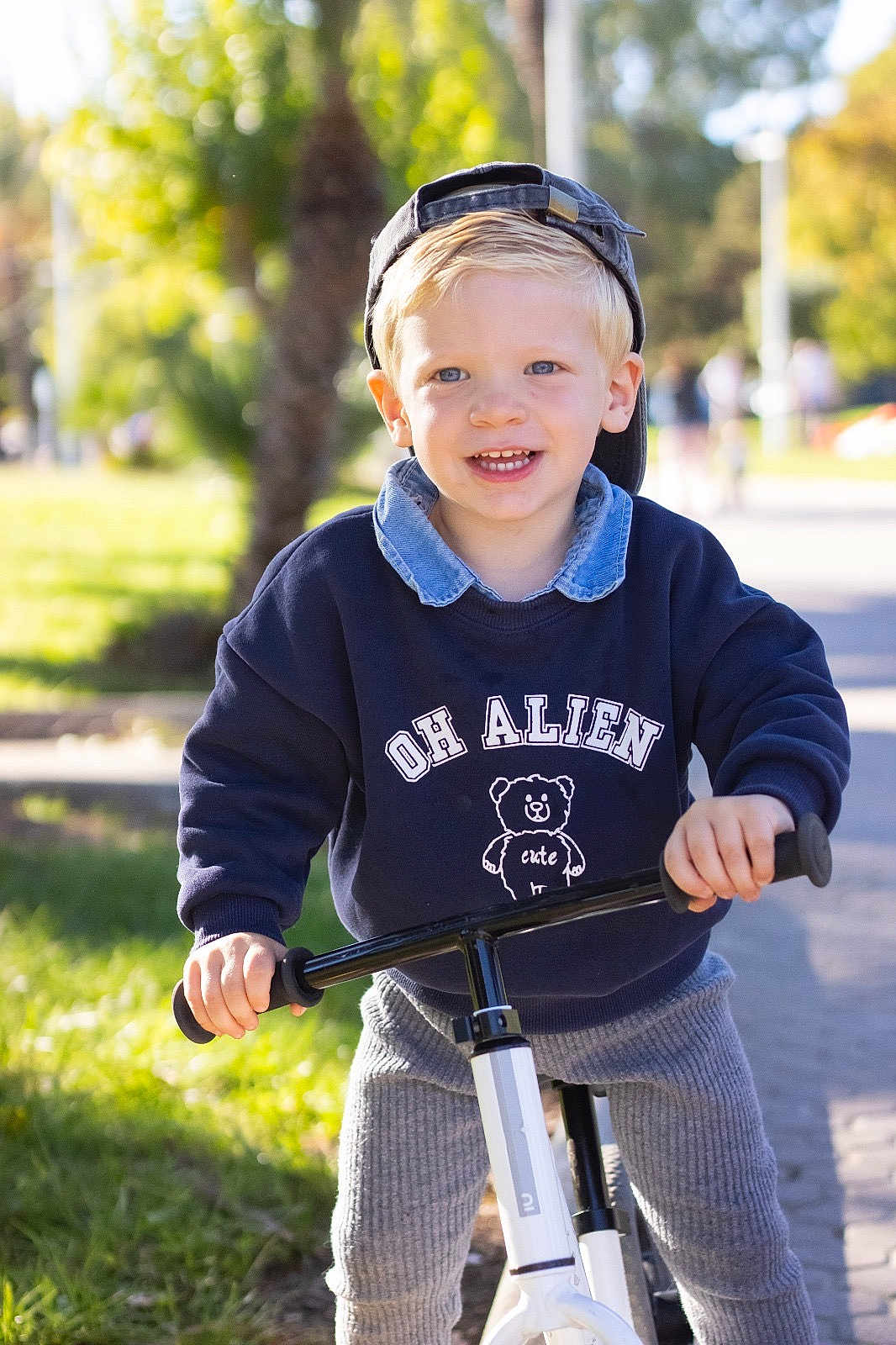 Giovanni a rejoint le concours — aidez-le/la à gagner de superbes lots ! toddler, child, bike, outdoor, daylight, park, sweatshirt, cap, smiling, blonde, cute, happy, play, casual_clothing, pants, young_child, person, greenery, sunny, recreation
