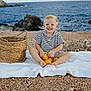 toddler, child, smiling, blonde_hair, striped_clothing, lemons, basket, beach, rocky_shore, blanket, sea, outdoor, happy, sitting, nature, coast, summer, vacation, playful, sunlight