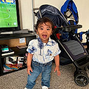 Favio is registered to the contest to win money with this photo: toddler, child, indoor, standing, jeans, shirt, sneakers, stroller, tv, soccer, carpet, furniture, hair, expression, room, toy, shelf, person, home, curly_hair