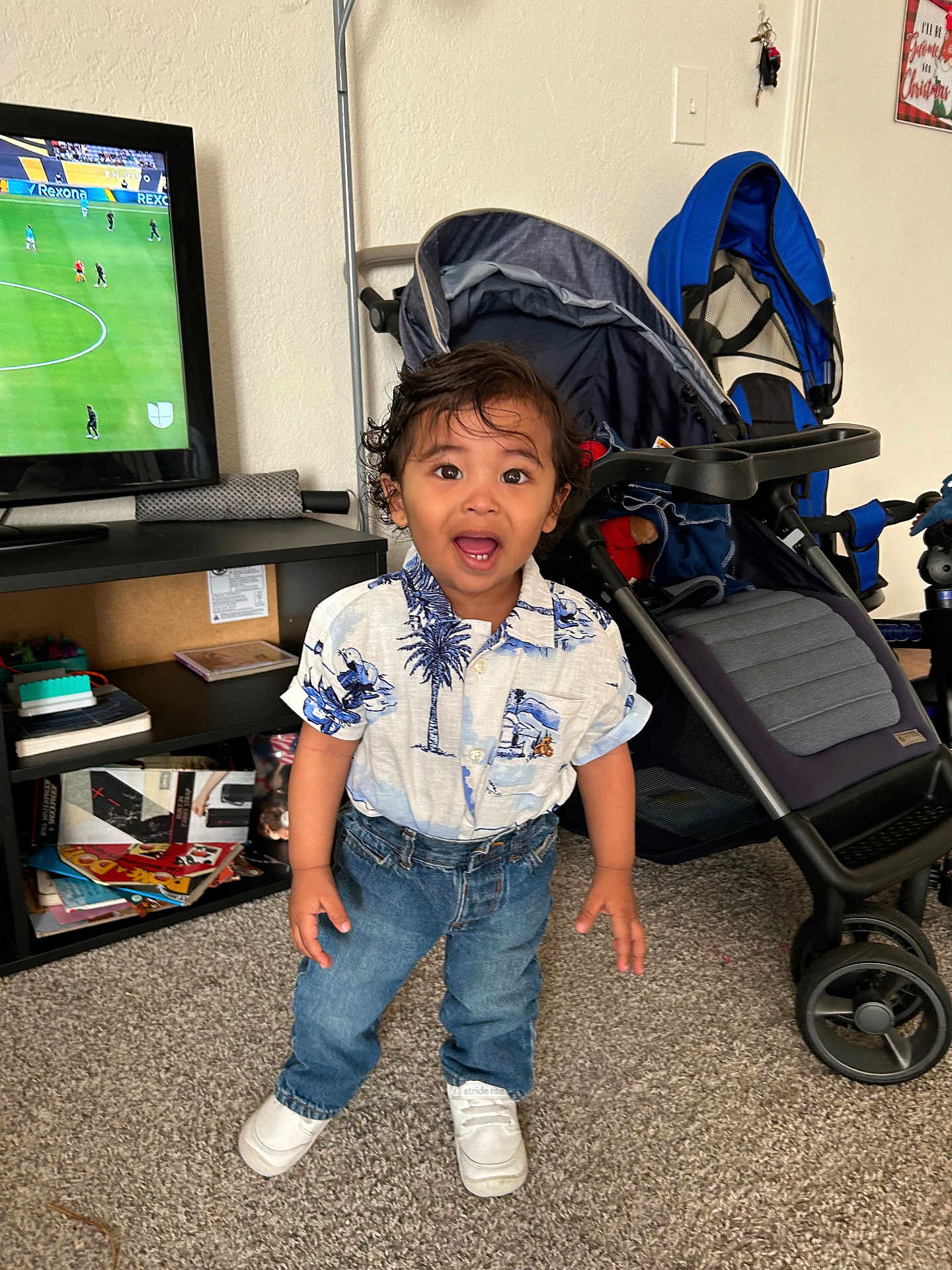 Favio is registered to the contest to win money with this photo: toddler, child, indoor, standing, jeans, shirt, sneakers, stroller, tv, soccer, carpet, furniture, hair, expression, room, toy, shelf, person, home, curly_hair