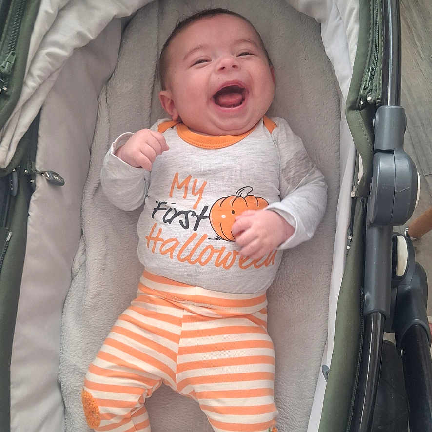 Rio is registered to the contest to win money with this photo: baby, celebration, child, clothing, cushion, cute, fabric, halloween, happy, holiday, indoors, infant, laughing, orange, person, pumpkin, smiling, striped_pants, stroller, white