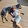 black_dog, boy, casual_clothing, child, daylight, dog, friendship, happy, nature, outdoor, pet, playful, relaxing, resting, snack, sneakers, summer, sunlight, tan_dog, wooden_deck