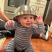 Maverick joined the competition — help win amazing prizes! baby, child, striped_onesie, metal_bowl, hat, smiling, happy, floor, kitchen, wooden_floor, adult_legs, sneakers, cup, brush, home, indoor, playful, person, cooking_tools, cute