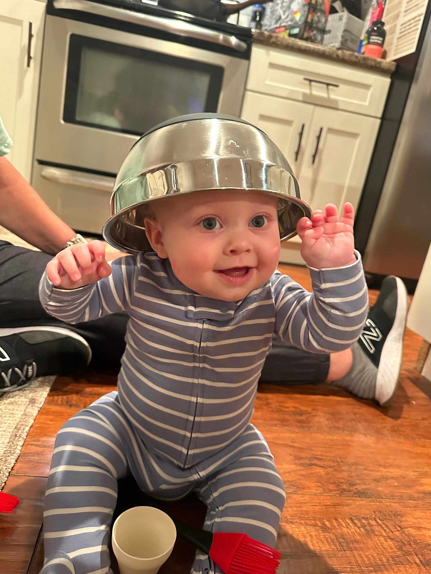 Maverick joined the competition — help win amazing prizes! baby, child, striped_onesie, metal_bowl, hat, smiling, happy, floor, kitchen, wooden_floor, adult_legs, sneakers, cup, brush, home, indoor, playful, person, cooking_tools, cute