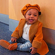 Javonte is registered to the contest to win money with this photo: baby, child, smile, orange_coat, beret, jeans, shoes, floor, carpet, wooden_wall, jewelry, happy, sitting, cute, indoor, portrait, person, fashion, accessory, clothing