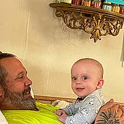 Asta is registered to the contest to win money with this photo: baby, man, indoor, holding, smiling, beard, tattoo, pajamas, shelf, jars, mirror, wall, person, clothing, face, expression, cute, curious, home, furniture