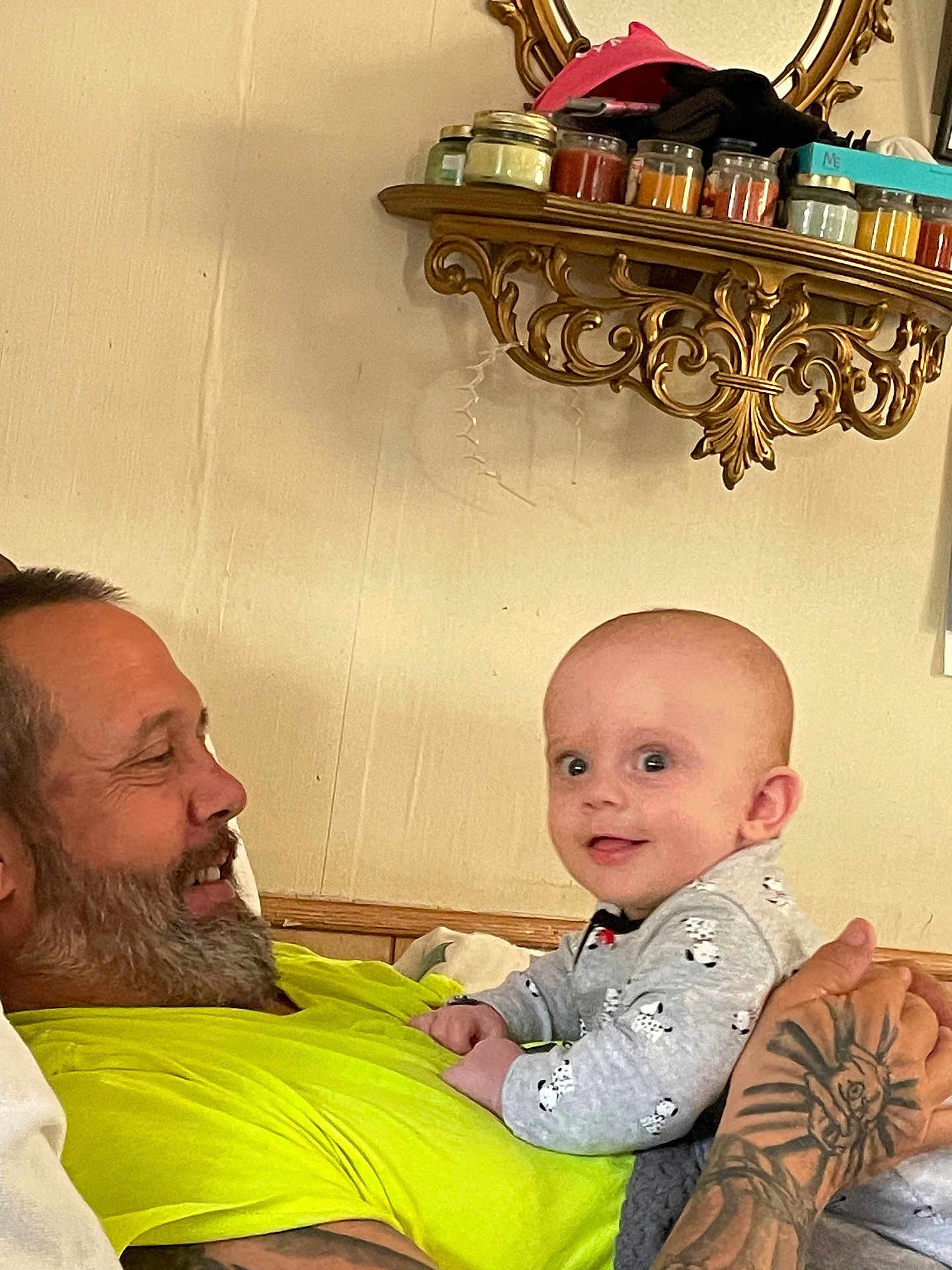 Asta is registered to the contest to win money with this photo: baby, man, indoor, holding, smiling, beard, tattoo, pajamas, shelf, jars, mirror, wall, person, clothing, face, expression, cute, curious, home, furniture