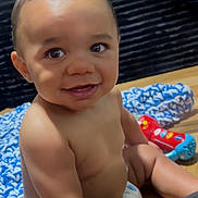 Ahlija is registered to the contest to win money with this photo: baby, infant, child, smiling, sitting, diaper, portrait, indoors, wooden_floor, knitted_blanket, colorful_toy, toy, dark_sofa, happy, bare_chested, curly_hair, face, close_up, photography, playtime