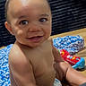 baby, infant, child, smiling, sitting, diaper, portrait, indoors, wooden_floor, knitted_blanket, colorful_toy, toy, dark_sofa, happy, bare_chested, curly_hair, face, close_up, photography, playtime