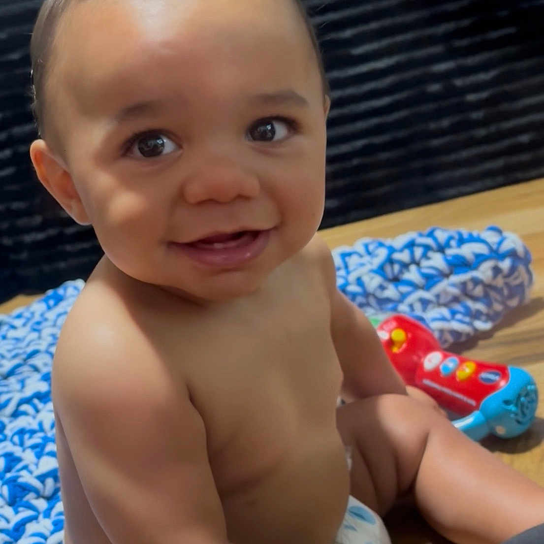 Ahlija is registered to the contest to win money with this photo: baby, infant, child, smiling, sitting, diaper, portrait, indoors, wooden_floor, knitted_blanket, colorful_toy, toy, dark_sofa, happy, bare_chested, curly_hair, face, close_up, photography, playtime