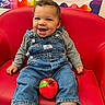 baby, child, infant, smiling, happy, portrait, sitting, red_chair, denim_overalls, gray_shirt, white_socks, toy, apple_toy, classroom, colorful_background, indoor, furniture, face, curly_hair, photography