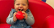 Adrian is registered to the contest to win money with this photo: baby, child, sitting, red_chair, toy, smiling, happy, face, portrait, classroom, colorful, artwork, rainbow, overalls, denim, long_sleeves, indoor, playroom, chewing, joyful