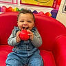 baby, child, sitting, red_chair, toy, smiling, happy, face, portrait, classroom, colorful, artwork, rainbow, overalls, denim, long_sleeves, indoor, playroom, chewing, joyful