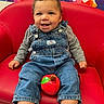 baby, child, smiling, sitting, portrait, indoor, red_couch, denim_overalls, gray_shirt, white_socks, toy_apple, colorful_background, classroom, furniture, happy, face, arms, legs, closeup, photography