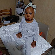 Maëna participe au concours pour gagner de l'argent avec cette photo : toddler, child, headband, cushion, table, indoor, person, clothing, socks, face, hand, wooden_chair, photographs, wall, room, furniture, curious, seated, baby, head
