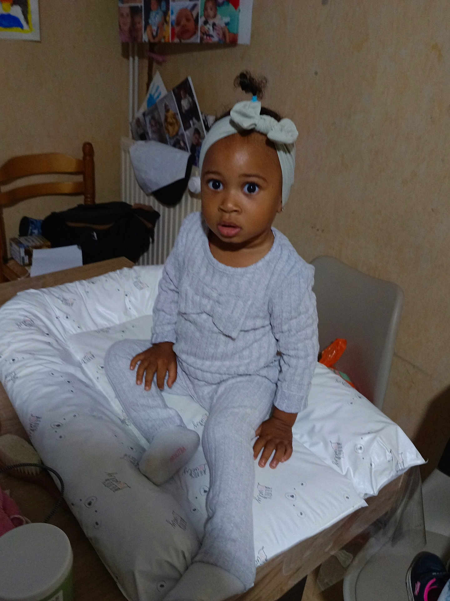 Maëna participe au concours pour gagner de l'argent avec cette photo : toddler, child, headband, cushion, table, indoor, person, clothing, socks, face, hand, wooden_chair, photographs, wall, room, furniture, curious, seated, baby, head