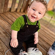 Jax joined the competition — help win amazing prizes! baby, child, smiling, sitting, curly_hair, blue_eyes, green_shirt, black_overalls, white_shoes, wooden_deck, outdoor, happy, cute, portrait, person, footwear, casual_clothing, infant, daylight, playful