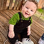 baby, child, smiling, sitting, curly_hair, blue_eyes, green_shirt, black_overalls, white_shoes, wooden_deck, outdoor, happy, cute, portrait, person, footwear, casual_clothing, infant, daylight, playful