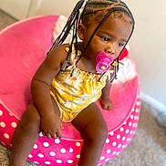 Ari is registered to the contest to win money with this photo: toddler, child, pacifier, braided_hair, yellow_outfit, pink_chair, polka_dots, indoor, carpet, seated, cute, young_child, portrait, baby, furniture, person, skin, head, expression, small_hand