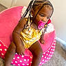 toddler, child, pacifier, braided_hair, yellow_outfit, pink_chair, polka_dots, indoor, carpet, seated, cute, young_child, portrait, baby, furniture, person, skin, head, expression, small_hand