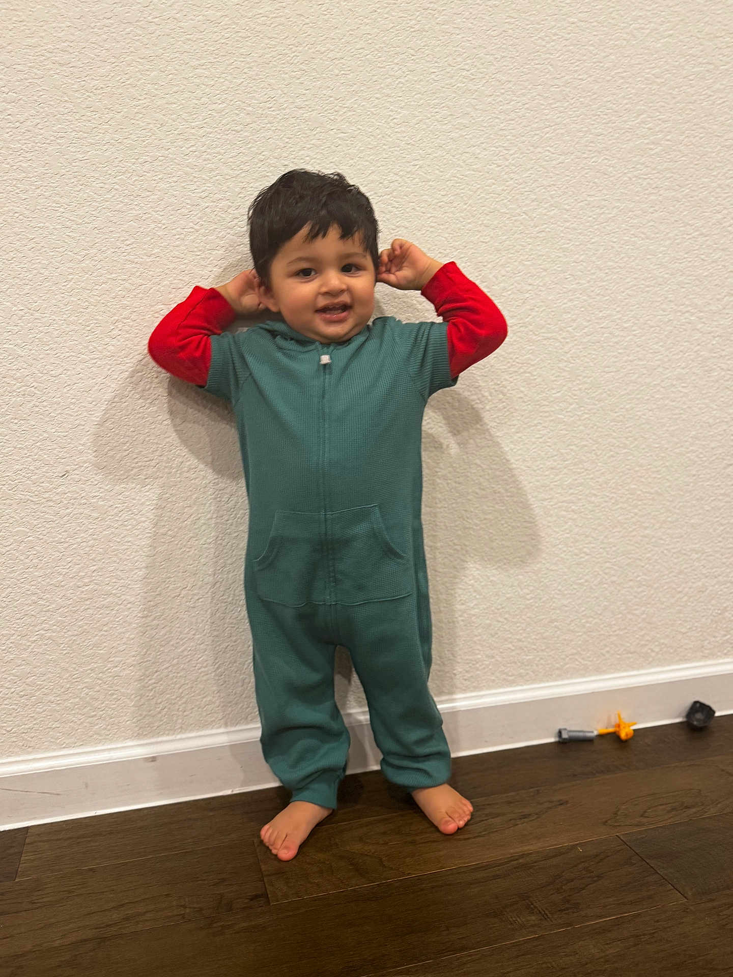 Shajun is registered to the contest to win money with this photo: toddler, child, onesie, green_clothing, red_sleeves, barefoot, standing, white_wall, wooden_floor, toys, playful, smiling, happy, indoor, person, young_child, cute, casual_clothing, home, portrait