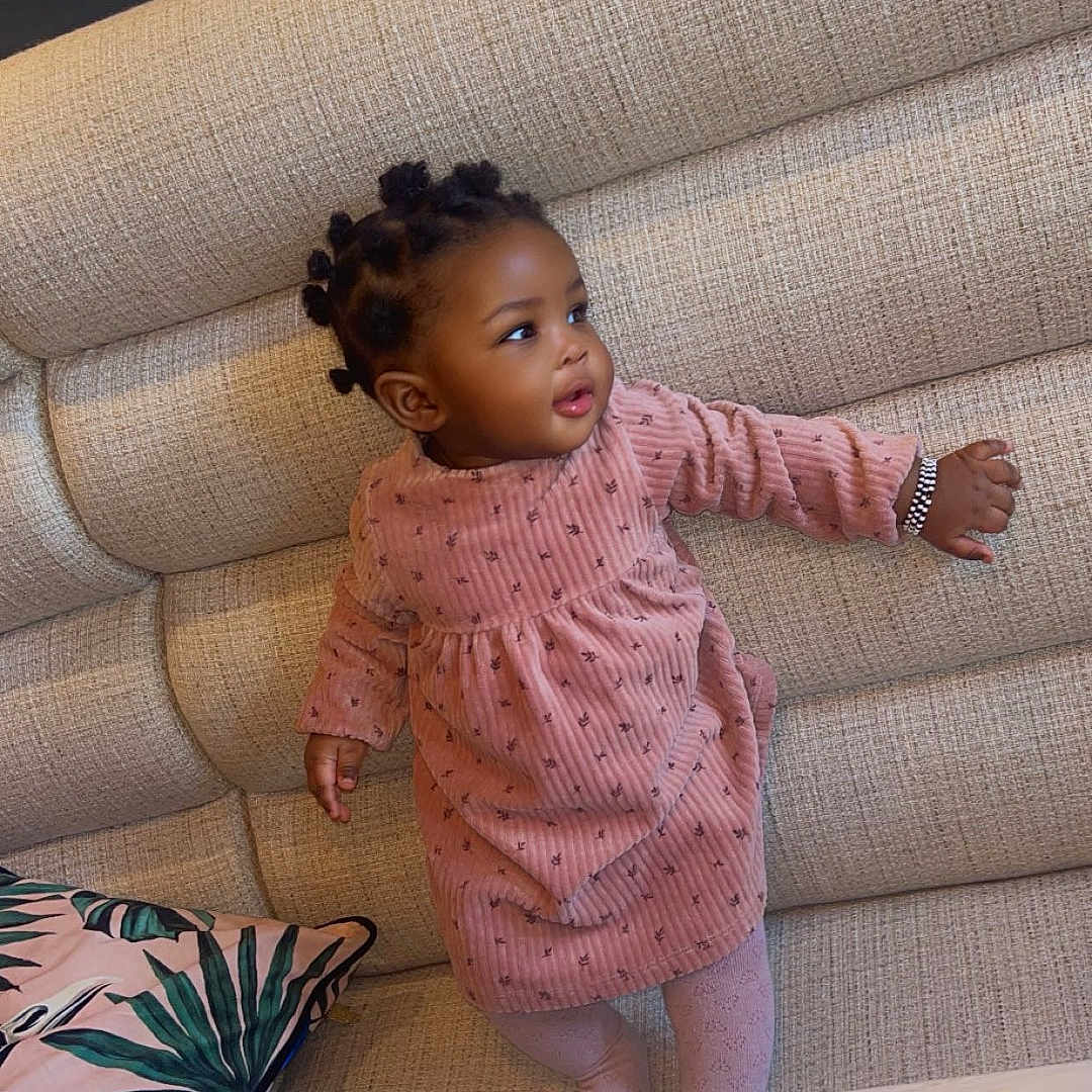 Hadja Mamah a rejoint le concours — aidez-le/la à gagner de superbes lots ! toddler, child, pink_dress, couch, cushion, pillow, table, bowl, tissues, indoor, person, cute, curious, standing, hair_buns, furniture, home, cozy, portrait, young_child