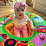 animal_print, baby, blue_eyes, cardboard_box, chair, child, colorful, curious_expression, daylight, floor, furniture, hand, headband, home, indoors, pink_clothing, play_mat, sitting, soft_toys, wooden_floor