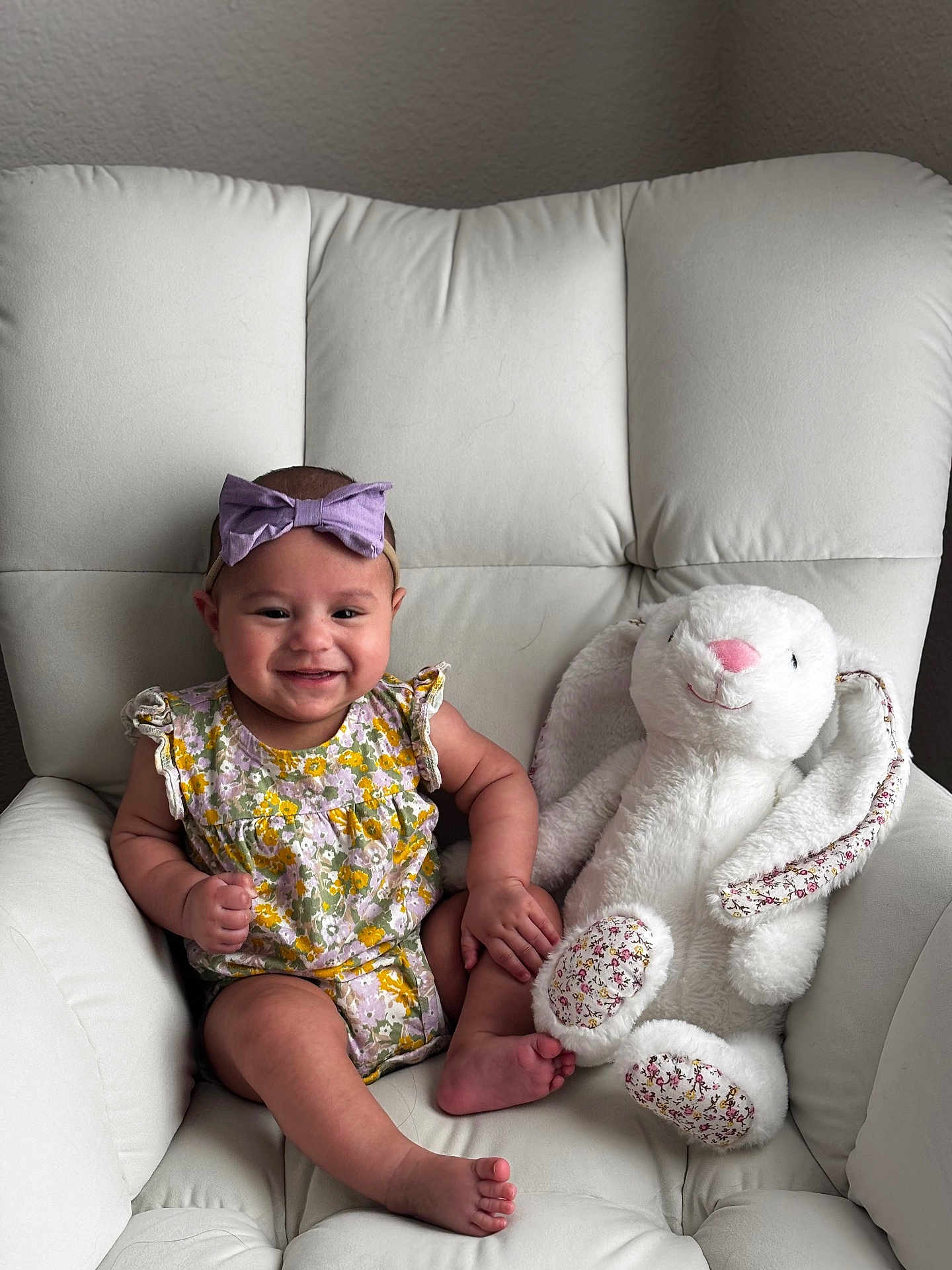 Luna is registered to the contest to win money with this photo: baby, child, smiling, purple_bow, floral_outfit, stuffed_bunny, white_chair, cushion, indoors, cute, happy, toy, infant, portrait, sitting, soft_toy, playful, headband, baby_boy, comfort