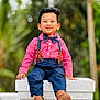 Diyan is registered to the contest to win money with this photo: child, boy, smiling, red_shirt, bow_tie, suspenders, blue_pants, brown_boots, outdoor, greenery, bokeh, portrait, sitting, fashion, cute, happy, face, hair, casual, daylight