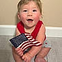 baby, child, flag, american_flag, red_shirt, gray_shorts, sitting, floor, wooden_floor, smiling, happy, blue_eyes, rosy_cheeks, indoor, person, one_person, cute, patriotic, young_child, caucasian