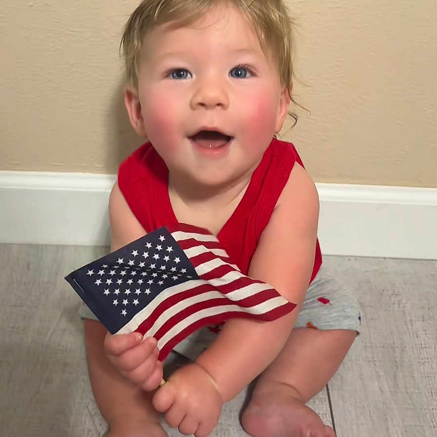 Tyler joined the competition — help win amazing prizes! american_flag, baby, blue_eyes, caucasian, child, cute, flag, floor, gray_shorts, happy, indoor, one_person, patriotic, person, red_shirt, rosy_cheeks, sitting, smiling, wooden_floor, young_child