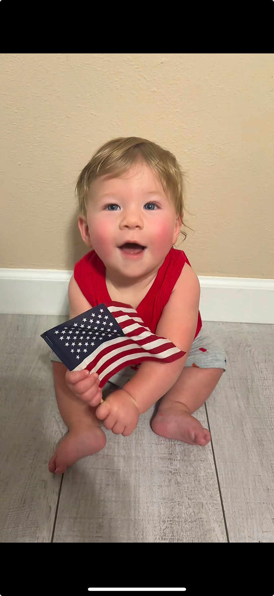 Tyler joined the competition — help win amazing prizes! baby, child, flag, american_flag, red_shirt, gray_shorts, sitting, floor, wooden_floor, smiling, happy, blue_eyes, rosy_cheeks, indoor, person, one_person, cute, patriotic, young_child, caucasian