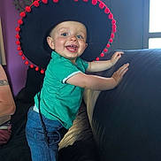 Gabin a rejoint le concours — aidez-le/la à gagner de superbes lots ! blue_eyes, child, clock, couch, decor, denim_shorts, face, furniture, green_shirt, hand, happy, hat, indoor, light, person, purple_wall, smiling, sombrero, toddler, window