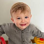 Gianni a rejoint le concours — aidez-le/la à gagner de superbes lots ! baby_toy, blond_hair, blue_eyes, button_up_shirt, cheeks, child, closeup, face, gray_shirt, hands, happy, indoor, looking_up, neutral_background, portrait, smiling, soft_lighting, steering_wheel, toddler, toy