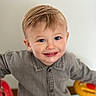 baby_toy, blond_hair, blue_eyes, button_up_shirt, cheeks, child, closeup, face, gray_shirt, hands, happy, indoor, looking_up, neutral_background, portrait, smiling, soft_lighting, steering_wheel, toddler, toy