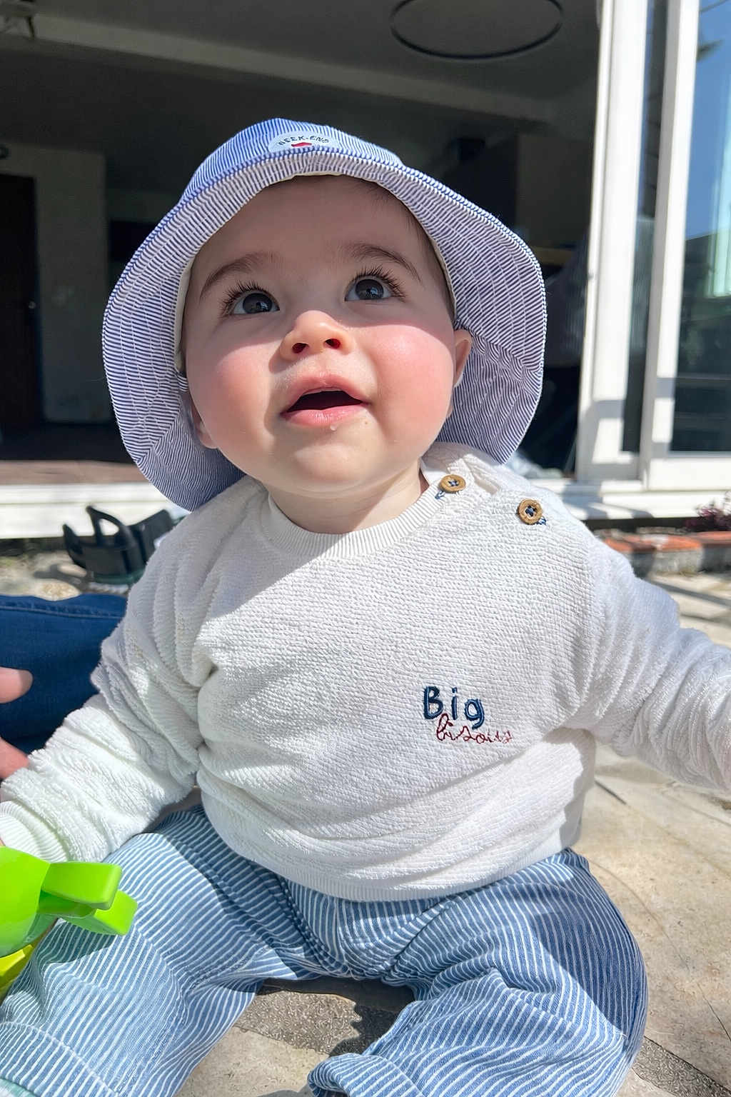 Achille a rejoint le concours — aidez-le/la à gagner de superbes lots ! baby, infant, bucket_hat, striped_hat, blue_and_white, big_eyes, long_eyelashes, white_sweater, striped_pants, green_toy, sitting, outdoor, sunlight, portrait, cheeks, cute, buttons, patio, window, candid