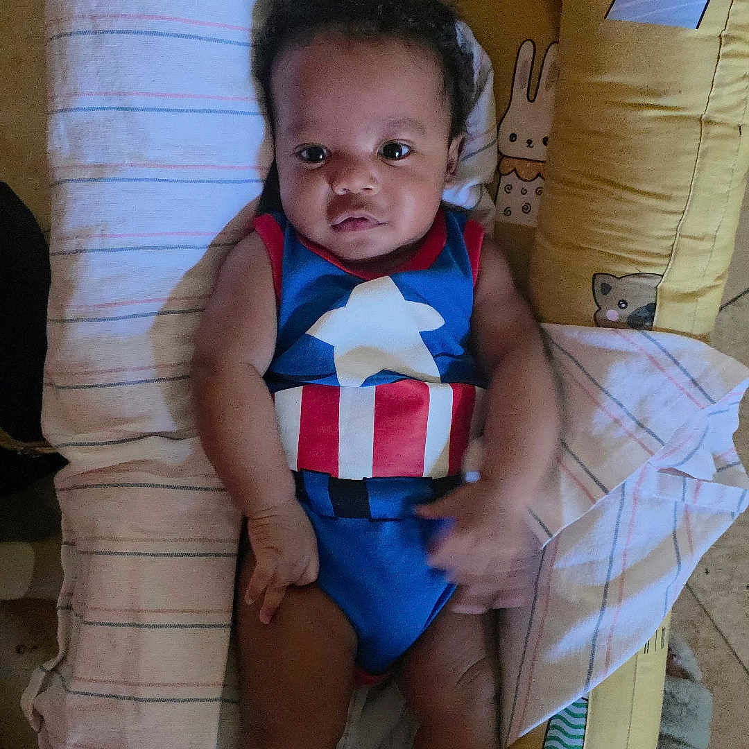 Aerrrow is registered to the contest to win money with this photo: baby, child, infant, onesie, superhero_costume, captain_america, yellow_cushion, animal_print, black_socks, face, eyes, hands, legs, person, indoor, floor, blanket, striped_cloth, cute, portrait