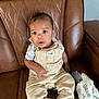 baby, child, overalls, beige_clothing, white_shirt, socks, brown_leather_chair, plush_toy, blanket, indoor, seated, curious_expression, wide_eyes, infant, portrait, furniture, soft_toys, comfortable, casual, home