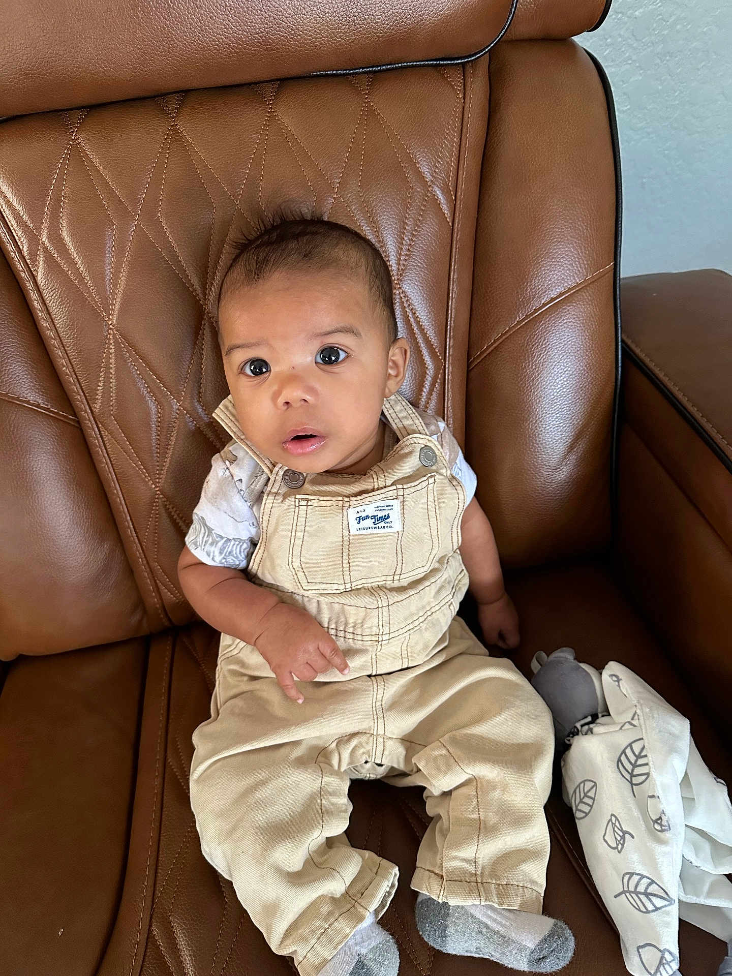 Zaire joined the competition — help win amazing prizes! baby, child, overalls, beige_clothing, white_shirt, socks, brown_leather_chair, plush_toy, blanket, indoor, seated, curious_expression, wide_eyes, infant, portrait, furniture, soft_toys, comfortable, casual, home