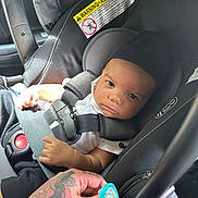 Zaire is registered to the contest to win money with this photo: baby, car_seat, infant, pacifier, hand, tattoo, seatbelt, car_interior, child, safety_harness, person, clothing, black_clothing, white_shirt, closeup, portrait, looking, indoor, young_child, seat