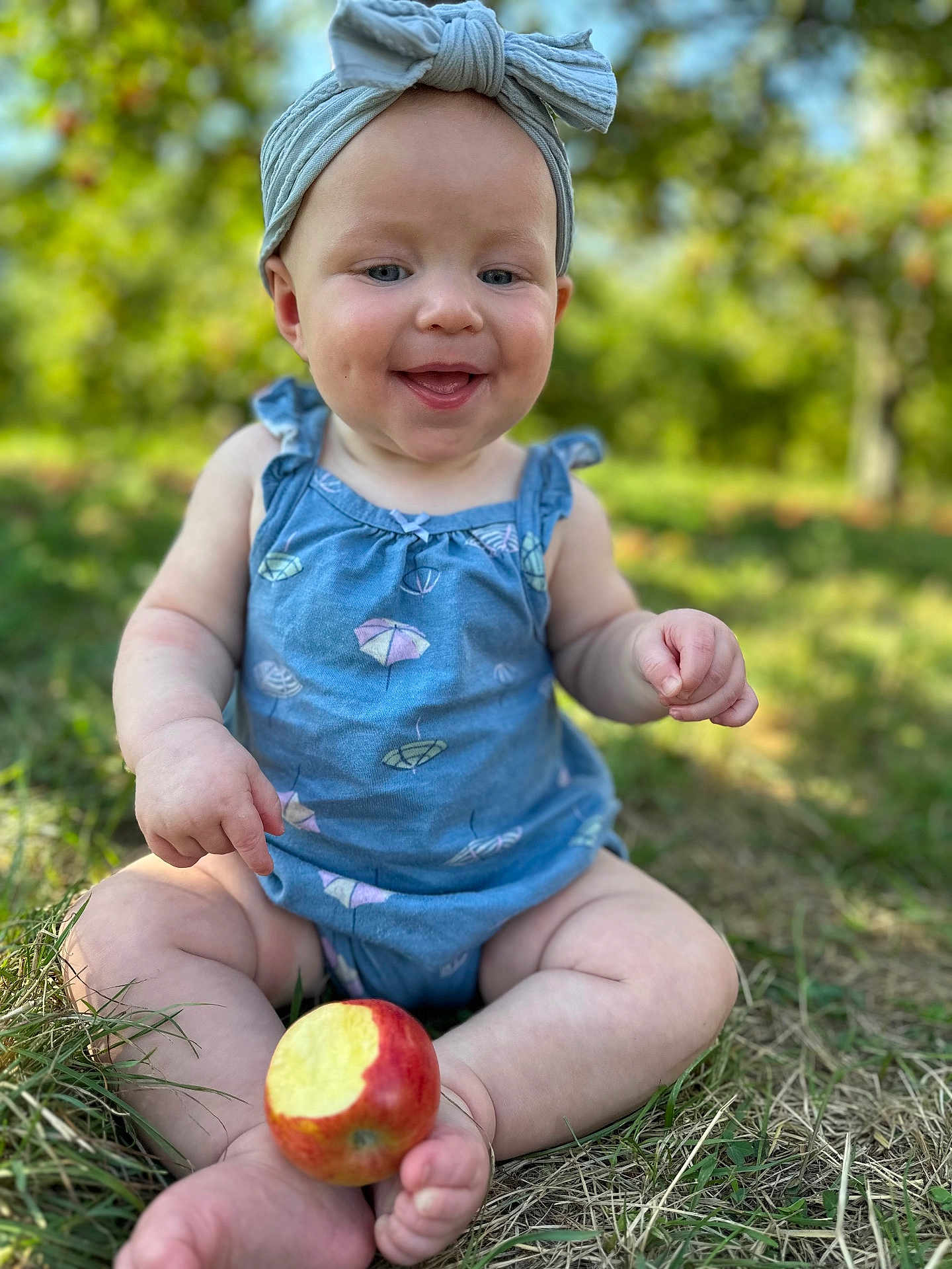 Olivia joined the competition — help win amazing prizes! baby, child, outdoor, grass, apple, bite, blue_clothing, head_wrap, smiling, happy, sitting, nature, greenery, summer, infant, closeup, cute, foot, hand, playful