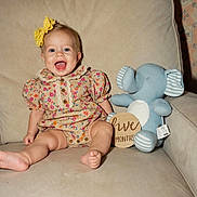 Eloise is registered to the contest to win money with this photo: baby, infant, child, sitting, smiling, laughing, portrait, indoor, couch, cushion, stuffed_animal, elephant_toy, floral_outfit, yellow_bow, baby_girl, six_months, milestone, wallpaper, home, cozy