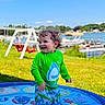 boats, child, curly_hair, daytime, fun, grass, green_shirt, happy, lake, outdoor, play, shark_design, smiling, splash_pad, summer, sunny, swimsuit, swing_set, toddler, water_play