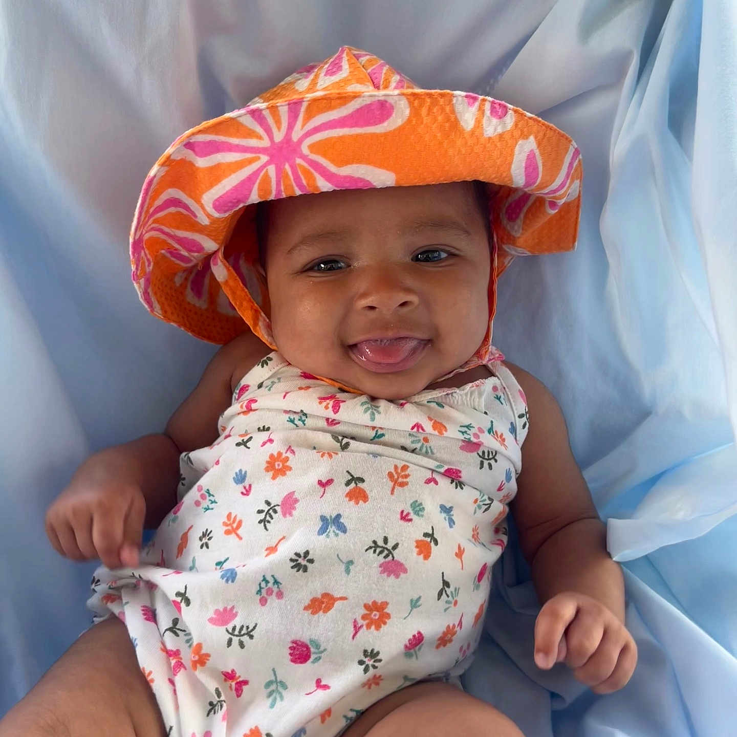 Tasia is registered to the contest to win money with this photo: baby, blue_background, child, clothing, cute, floral_pattern, hand, happy, hat, infant, leg, lying_down, onesie, person, playful, portrait, skin, smiling, sunhat, tongue_out