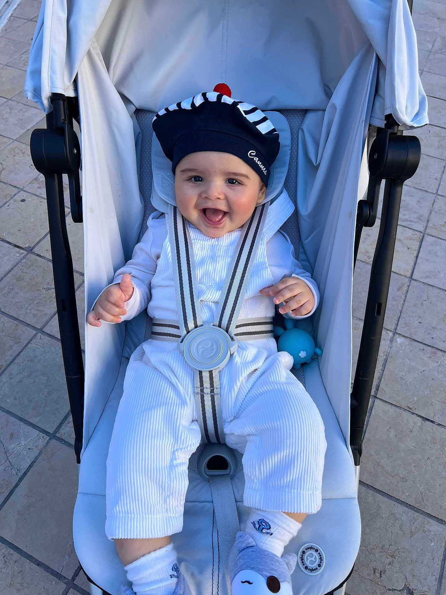 Gabriel participe au concours pour gagner de l'argent avec cette photo : baby, infant, stroller, harness, smiling, hat, bonnet, toy, outdoor, pavement, seat, happy, portrait, cute, clothing, socks, shoes, cheeks, hands, seatbelt