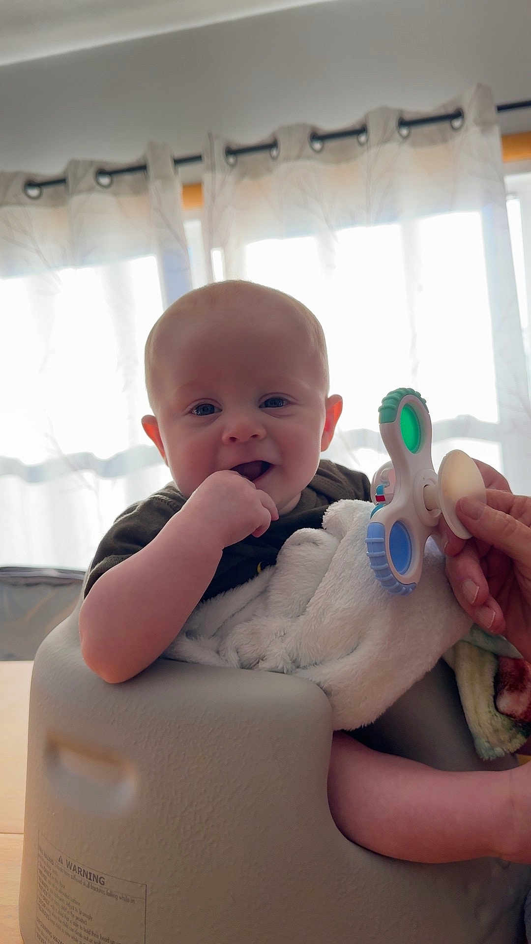 Braxton joined the competition — help win amazing prizes! baby, infant, smile, bald_head, hand_in_mouth, toy, fidget_spinner, blanket, baby_seat, curtains, backlight, parent_hand, indoor, cheeks, eyes, happy, portrait, furniture, table, soft_toy