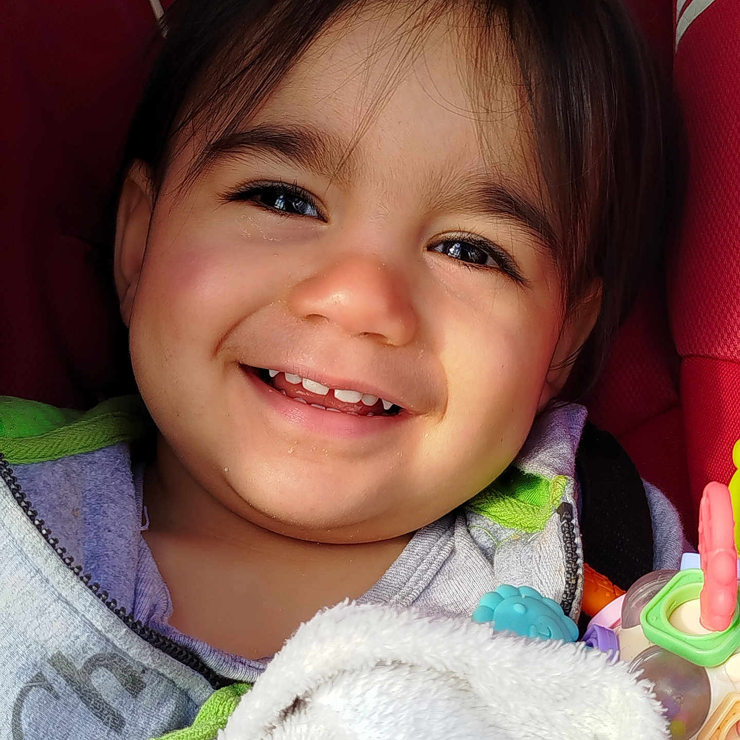 Orion participe au concours pour gagner de l'argent avec cette photo : blanket, candid, car_seat, cheeks, child, closeup, dark_hair, eyes, face, happy, hoodie, plush, portrait, rattle, red_seat, smile, sunlight, teeth, toddler, toy